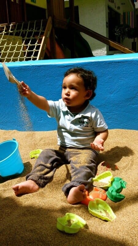 Niño pequeño lanza arena al aire mientras juega sentado en el arenero