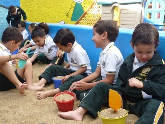 Grupo de alumnos en uniforme jugando descalzos con cubetas y palas en el arenero del colegio