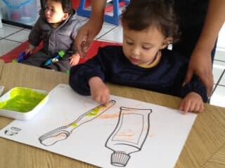 Niña pequeña colorea un dibujo de un cepillo y una pasta de dientes con ayuda de una maestra