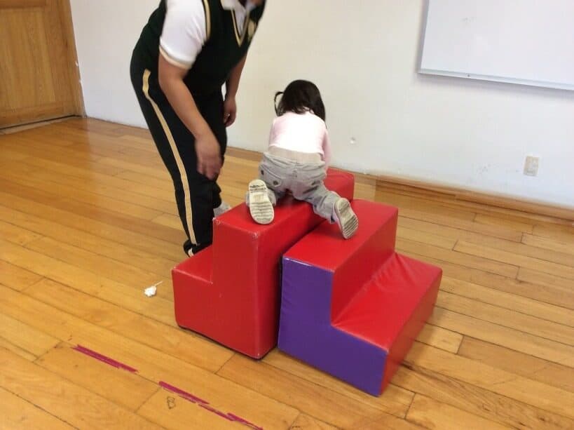 Niña pequeña sube el primer escalón de una rampa de espuma roja tomada de la mano de su maestra
