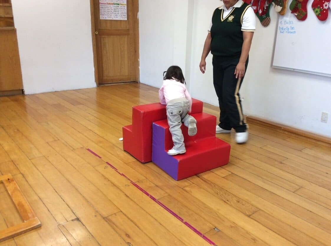 Niña pequeña sube sola la rampa de espuma mientras su maestra la vigila de cerca