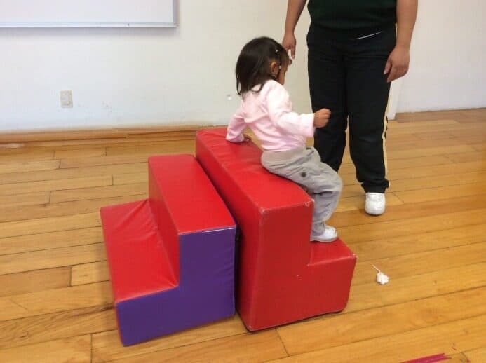 Niña pequeña se impulsa para subir la rampa de espuma sujetando la mano de su maestra