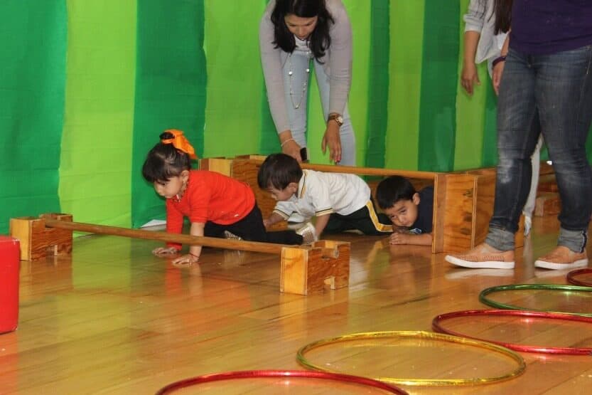 Niños pequeños gatean bajo unas vigas de madera entre aros de colores, acompañados por sus maestras
