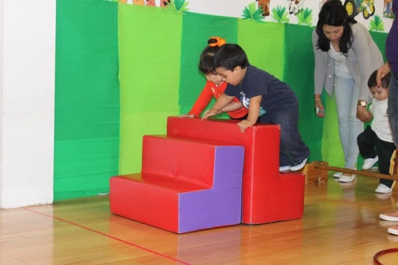 Niño pequeño trepa la rampa de espuma roja apoyado por su maestra, frente a un fondo verde
