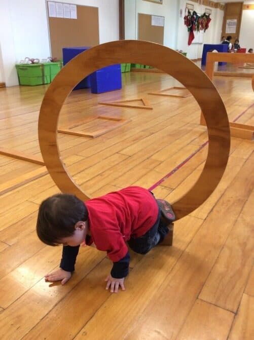 Niño con suéter rojo gatea bajo un aro grande de madera en el salón de psicomotricidad