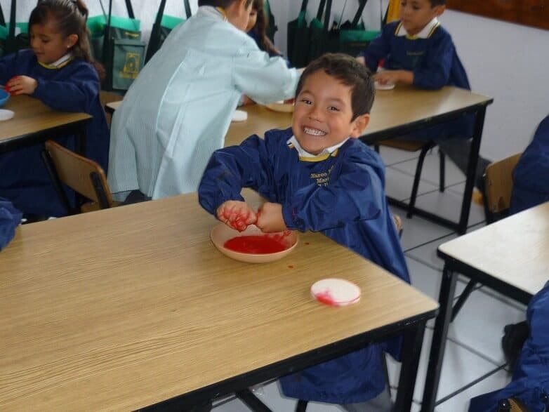 Alumno con bata azul sonríe en su mesa durante una actividad con pintura roja