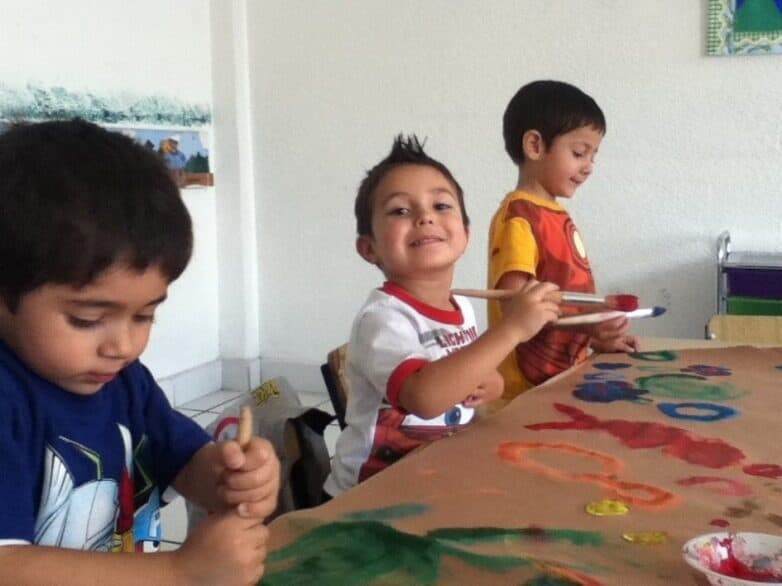 Tres alumnos pintan con pinceles sobre un pliego de papel compartido