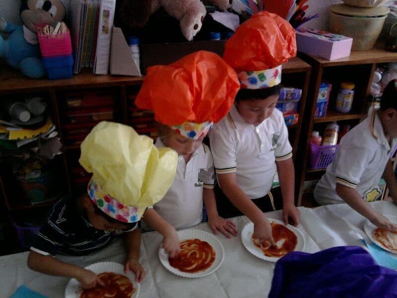 Alumnos con gorros de papel de colores untan salsa sobre sus bases de pizza