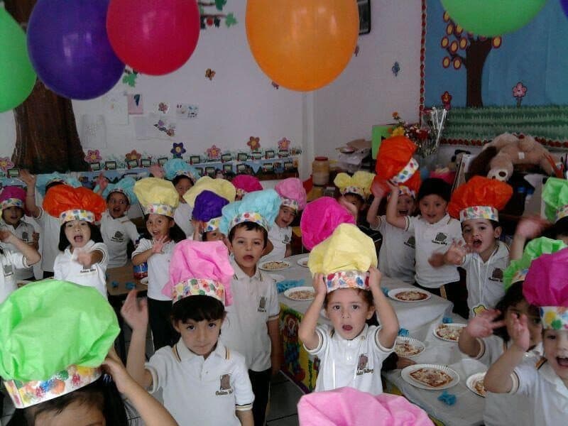 Grupo de alumnos con gorros de papel levantan la mano frente a sus platos, entre globos