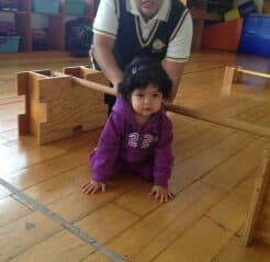 Niña pequeña gatea bajo una viga de madera baja acompañada por su maestra
