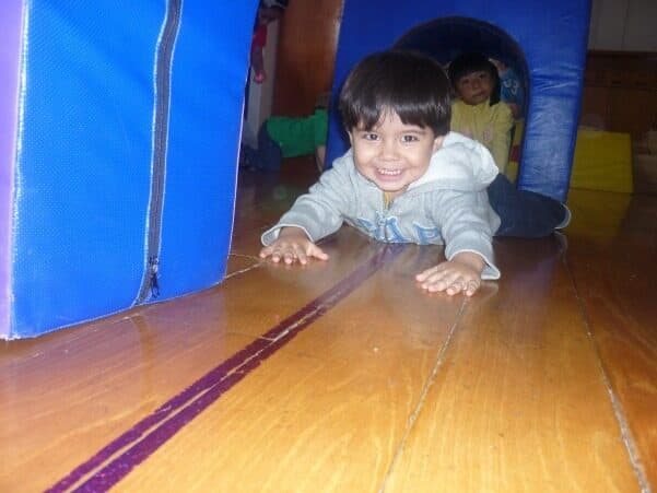 Niño pequeño sale gateando de un túnel de colchoneta azul, sonriendo a la cámara