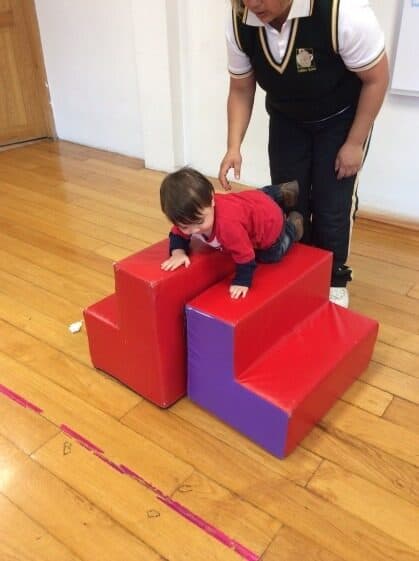 Niño pequeño sube el primer escalón de la rampa de espuma con apoyo de su maestro