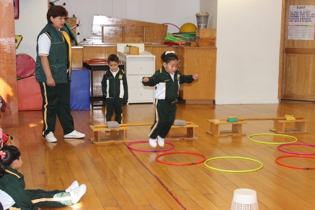 Alumnos saltan sobre bancos de madera y aros durante el circuito, guiados por su maestra